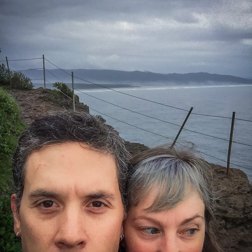 A man and a woman stand outdoors on a rocky path near a body of water with a cloudy sky in the background.