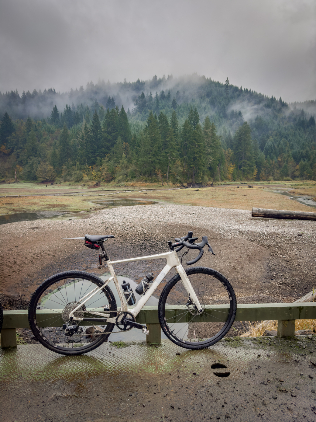 Photograph of a gravel bicycle on a bridge railing, the middle ground is a reservoir that is mostly empty of water, the background is forested with wisps of fog mixed in.