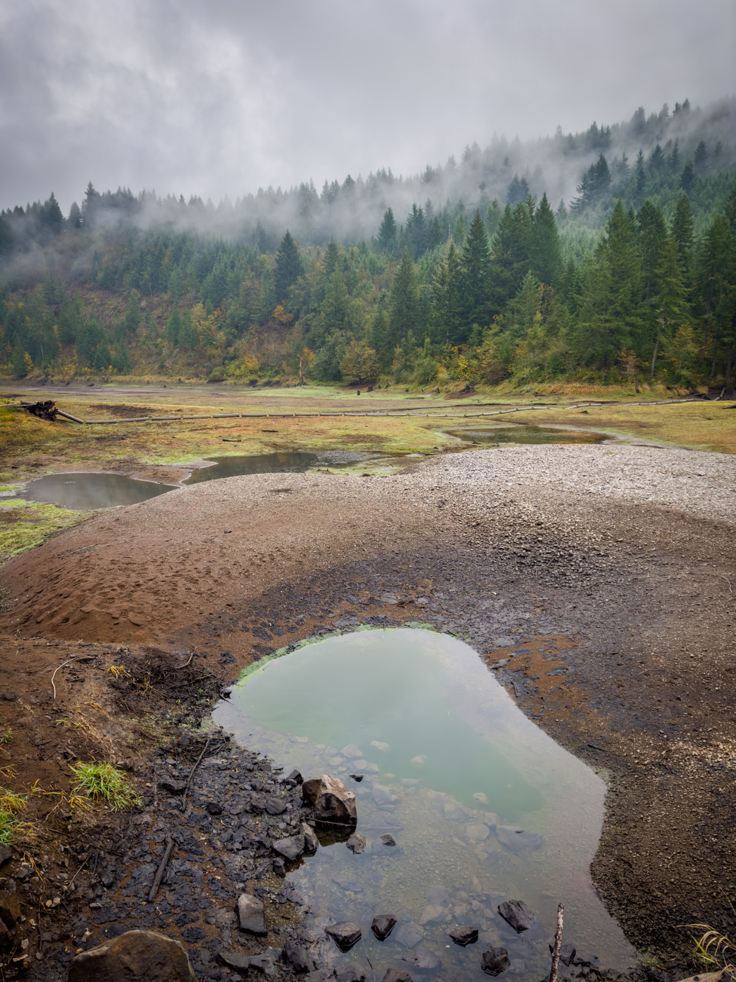 Photograph of a reservoir that is mostly empty of water, the background is forested with wisps of fog mixed in.
