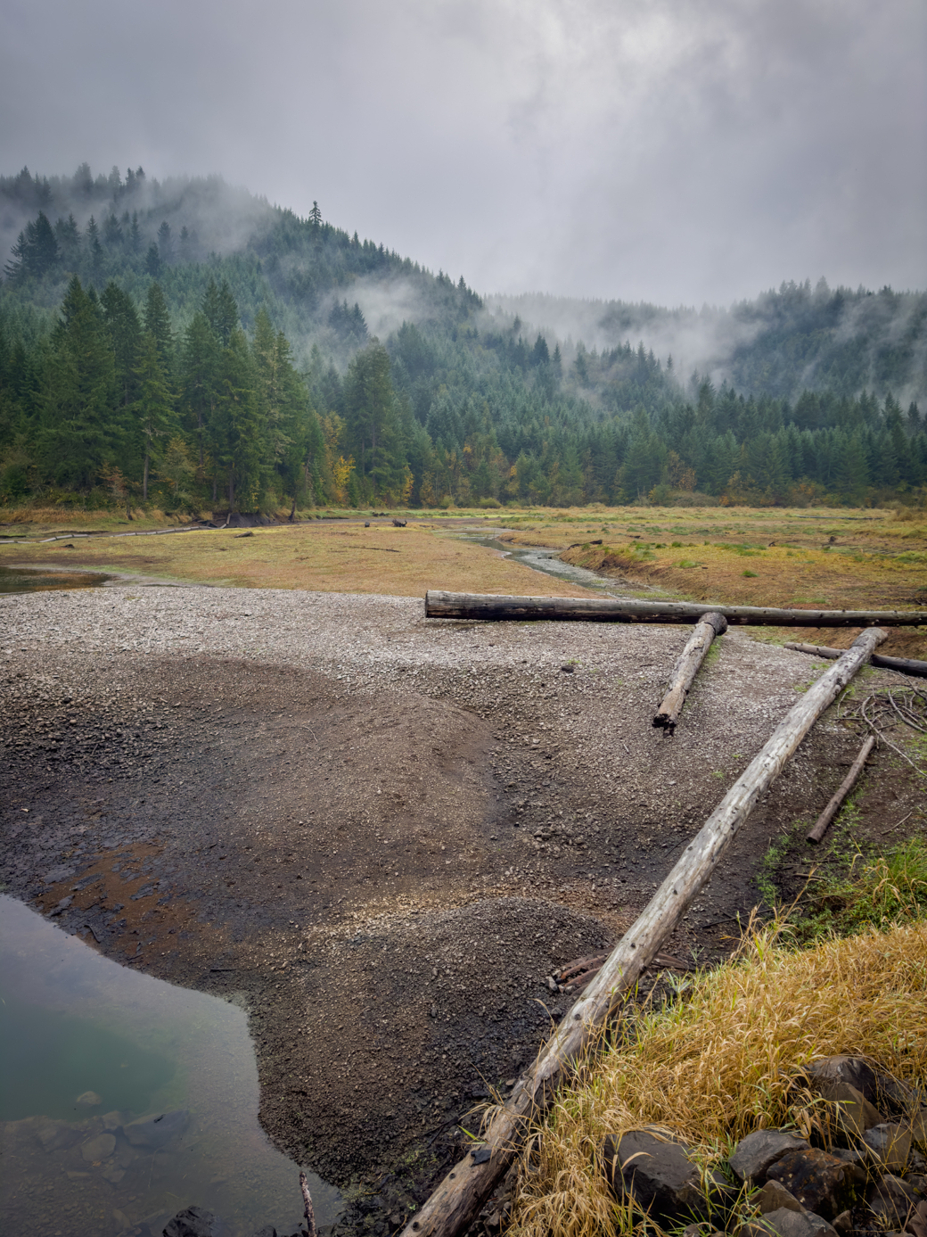 Photograph of a reservoir that is mostly empty of water, the background is forested with wisps of fog mixed in.