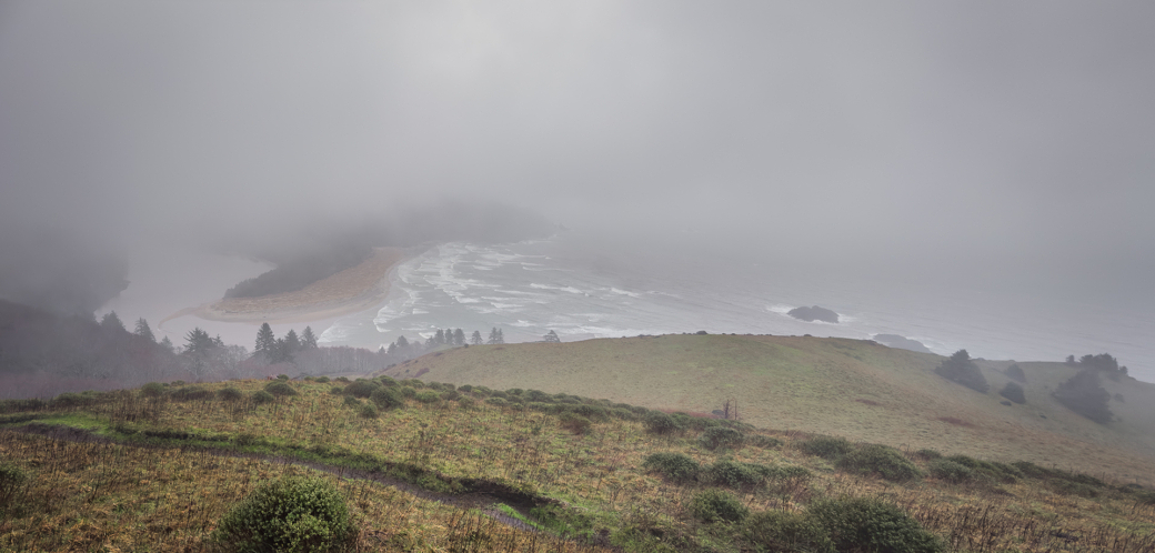 A foggy coastal landscape with rolling hills foregrounding a mist-covered beach and ocean.